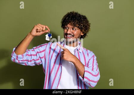 Photo of overjoyed guy with stubble wavy hairstyle dressed striped shirt directing look at keys in arm isolated on khaki color background Stock Photo