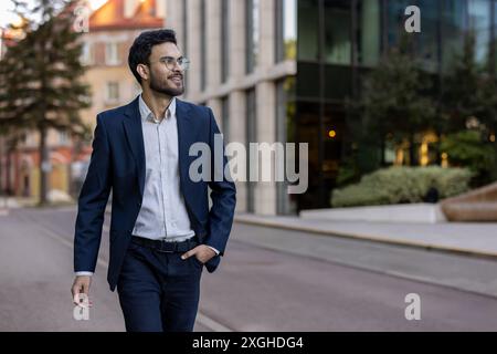 Confident smiling businessman wearing blazer and glasses walking in urban city street. Professional young man enjoying his day. Concept of success, confidence, business attire. Stock Photo