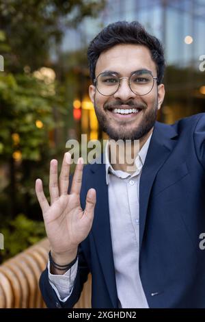 Professional businessman wearing glasses and blue suit smiling and waving. Video call concept, taken outside modern office building. Friendly, positive, confident business communication. Stock Photo