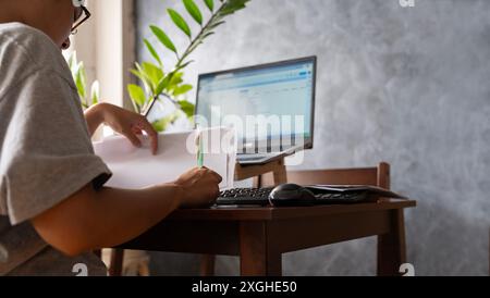 Woman working on laptop at home office while looking at documents in warm lighting atmosphere through the window. Stressed woman working from home at Stock Photo