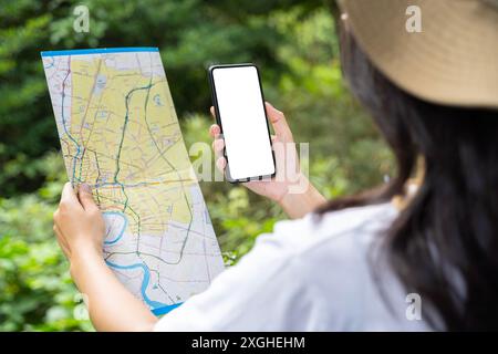 Close up of woman hands with a smartphone and map in the forest background, isolate screen. Happy young woman with a map, smartphone travelling on nat Stock Photo