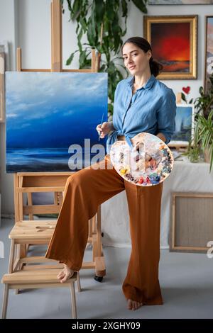 Confident artist with colour palette looks at camera stands near easel with painting in art studio. Stock Photo