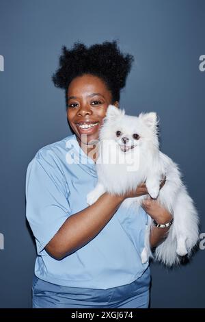 Vertical medium shot portrait of African American female vet expert in scrubs holding white puppy close smiling while looking at camera isolated on blue background in studio, camera flash Stock Photo