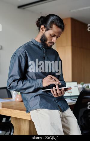 Young Indian man leaning on wooden table and using white tablet to find information in Internet Stock Photo