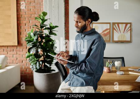 Young Indian entrepreneur checking his mail via tablet while leaning on wooden work desk in cozy and spacious office Stock Photo