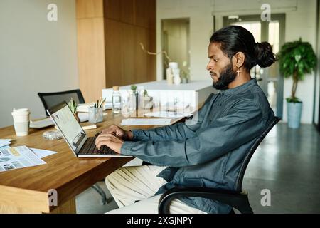 Highly qualified IT specialist sitting at wooden work table and writing analytical report using laptop in modern office fully equipped with office supplies and furniture Stock Photo