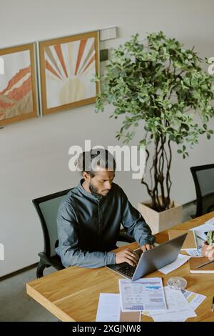 Young Indian businessman dressed in deep blue flax shirt sitting at wooden table and using silver laptop in spacious office Stock Photo