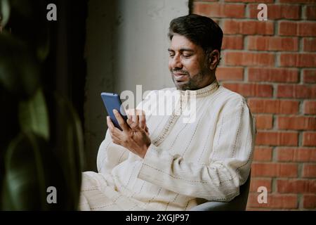 Middle aged Indian man communicating in social network and smiling while sitting in chair Stock Photo