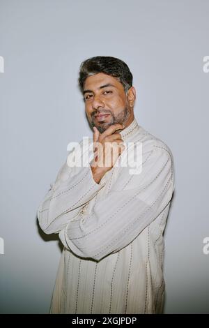Portrait of young Indian man dressed in patterned creamy shirt holding his chin and smirking with squint while sitting for photo Stock Photo