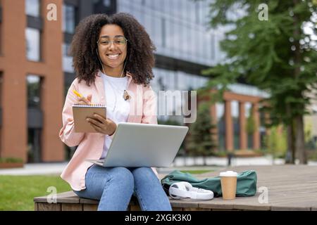 Smiling student with curly hair working on notes and laptop outdoors. Young woman sitting outside modern building with notepad and coffee, studying in pleasant environment Stock Photo