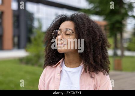 Young woman with curly hair and glasses stands peacefully outdoors in urban park. She breathes deeply, enjoying nature and tranquility. Capturing serene content moment in bustling city environment. Stock Photo