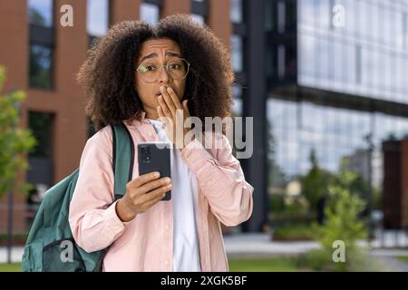 Young student with curly hair and glasses holding smartphone with shocked expression outdoor. Brick and glass building in background. Concept of surprise, technology, urban lifestyle. Stock Photo