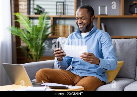 Smiling man working remotely from home, using smartphone and reviewing document while sitting on sofa with laptop and notepad. Concept of remote work home office, happiness, productivity, technology. Stock Photo
