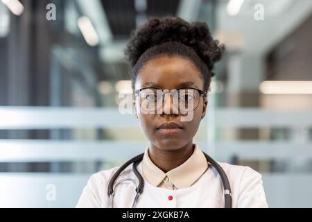 Female doctor wearing glasses and stethoscope standing confidently in modern medical office. Professional healthcare provider posing indoors, reflecting strength and competence Stock Photo