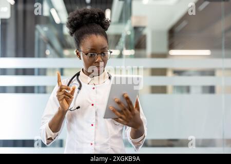 Female doctor wearing stethoscope using tablet for video call in modern office. Doctor making virtual consultation, communicating with patient. Professional healthcare worker using technology Stock Photo