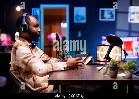 Side view portrait of african american male talk show host speaking with his online audience during a live podcast recording from his home studio, he wears wireless headphones. Stock Photo