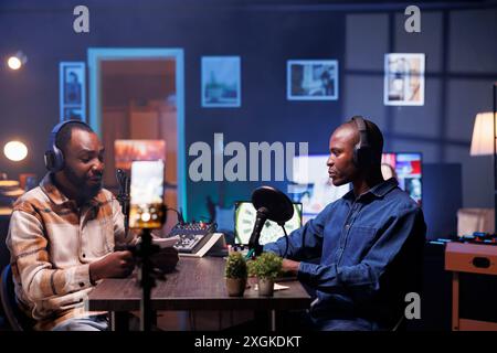 Vlogger is recording an interview with smartphone on tripod in studio while reviewing notes to ask quality questions to guest. Journalist using mobile device to film vlog while talking on mic with man Stock Photo