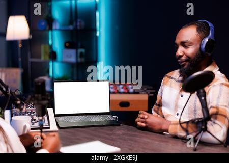 African man wears headset and speaks into mic while his laptop displays blank white screen. Device on desk shows an isolated chromakey mockup template while male content creator making a podcast. Stock Photo