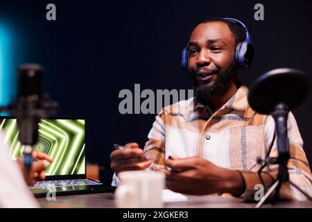 African american man wearing wireless headset and microphone nearby, is having a discussion with a person making a podcast. Male content creator using recording equipment to create a talk show. Stock Photo