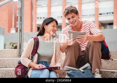Two teenagers sitting on the university campus stairs, studying lessons and doing homework using a tablet. Classmates sharing ideas for an academic project, working with a touchpad. College students. High quality photo Stock Photo