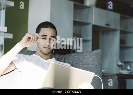 Young man relaxes at home, listening to music on a tablet with earphones while lounging on a sofa. Enjoying digital entertainment brings him peace and Stock Photo
