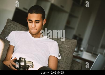 Young photographer reviewing photos on a couch, immersed in editing with a digital camera. Natural light highlights his passion for photography Stock Photo