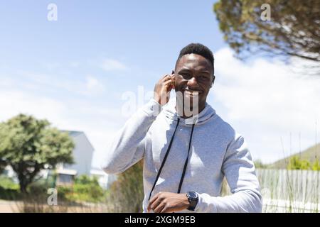 Smiling african american man wearing hoodie and earphones running in countryside. Jogging, fitness, exercise, wellbeing and healthy lifestyle, unalter Stock Photo