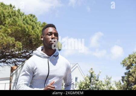 Happy african american man wearing hoodie and earphones running in countryside, copy space. Jogging, fitness, exercise, wellbeing and healthy lifestyl Stock Photo