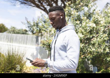 Focused african american man training outdoors, wearing earphones and using smartphone. Communication, jogging, fitness, exercise, wellbeing and healt Stock Photo