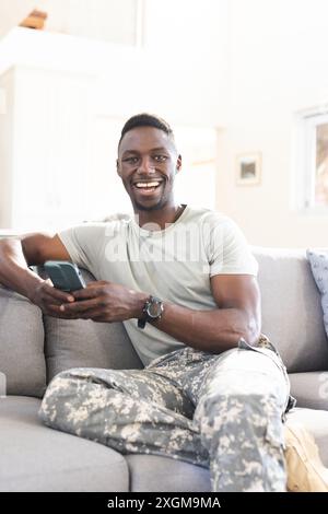 Portrait of happy african american male soldier on couch at home using smartphone, copy space. Communication, military service, army, return, home, do Stock Photo