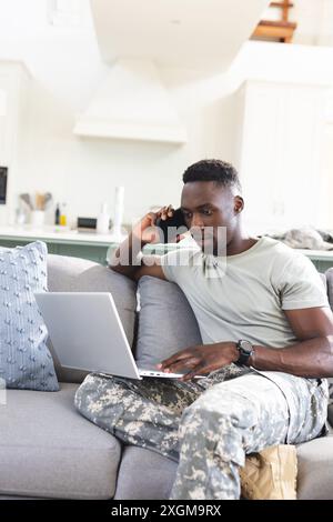 Focused african american male soldier at home using laptop and talking on smartphone, copy space. Communication, military service, army, return, home, Stock Photo