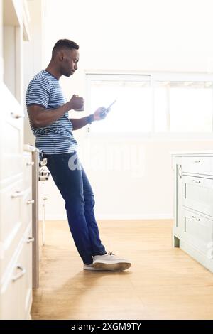 African american man drinking coffee and using smartphone standing in sunny kitchen, copy space. Communication, refreshment, morning, domestic life an Stock Photo