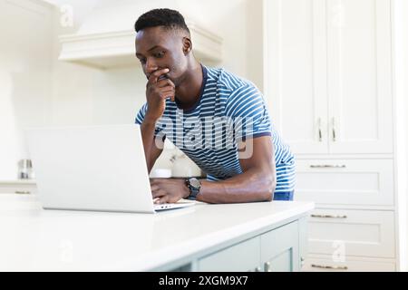 Thoughtful african american man using laptop leaning on worktop in sunny kitchen, copy space. Communication, refreshment, morning, domestic life and l Stock Photo