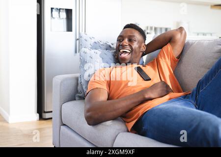Happy african american man relaxing on sofa laughing, watching tv, copy space. Relaxation, television, technology, entertainment, free time, domestic Stock Photo