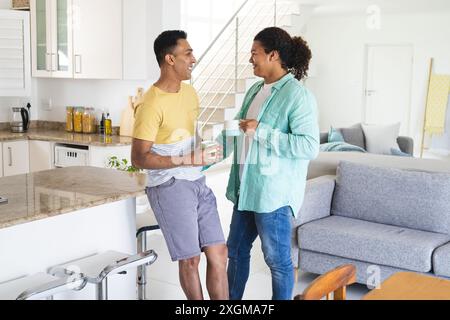 Middle Eastern man and young Biracial man, a gay couple, chatting in a bright home kitchen. They share a relaxed moment with coffee, reflecting a comf Stock Photo
