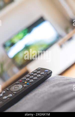 TV remote control in the foreground, tv in the blurry background. Streaming Stock Photo