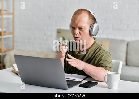 A man with inclusivity in casual clothing sits at a desk, wearing headphones and using a laptop. Stock Photo
