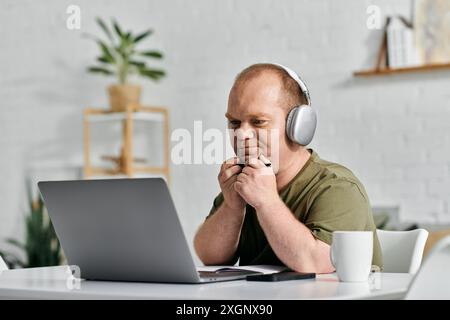 A man with inclusivity sits at a desk in his home office, working on his laptop while wearing headphones. Stock Photo