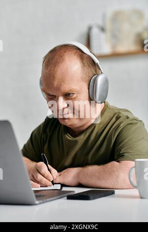 A man with inclusivity in a casual shirt sits at a desk with a laptop and takes notes while wearing headphones. Stock Photo