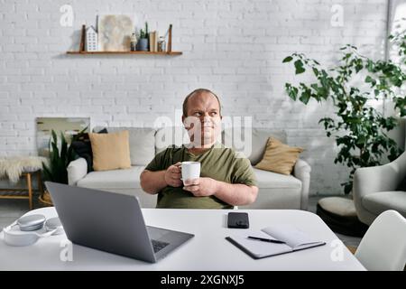 A man with inclusivity sits at a desk in a casual home office setting, holding a mug and gazing thoughtfully into the distance. Stock Photo