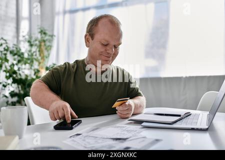 A man with inclusivity in casual attire uses his phone and credit card while seated at a home office desk. Stock Photo