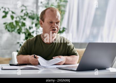 A man with inclusivity sits at a desk in a home office, reviewing documents while looking at a laptop. Stock Photo