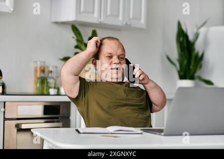 A man with inclusivity sits at his home office desk, talking on the phone, with a laptop and a notebook in front of him. Stock Photo