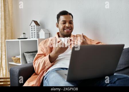 A man sits on a couch in his living room, using sign language to communicate while using a laptop. Stock Photo