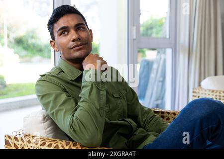 Young biracial man sits thoughtfully in a home setting, unaltered. He appears contemplative, relaxing on a wicker chair near a bright window. Stock Photo