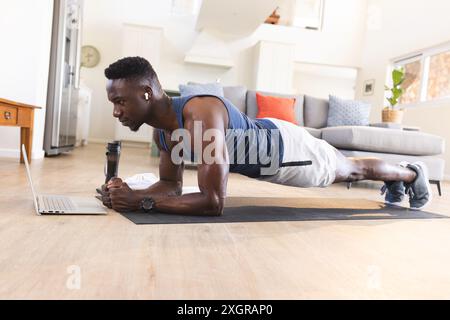 Fit african american man exercising at home, lying in plank position and using laptop. Communication technology, fitness, training, exercise, domestic Stock Photo