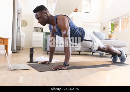 Fit african american man exercising at home, doing push ups and using laptop. Communication technology, fitness, training, exercise, domestic life and Stock Photo