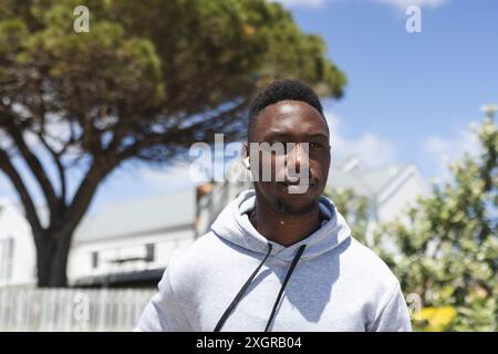 Focused african american man wearing hoodie and earphones running in sunny street. Jogging, fitness, exercise, wellbeing and healthy lifestyle, unalte Stock Photo