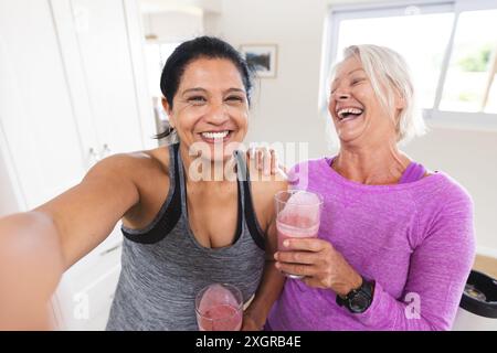 Two happy diverse senior female friends drinking fruit smoothies taking selfie in kitchen. Communication, technology, nutrition, friendship, domestic Stock Photo