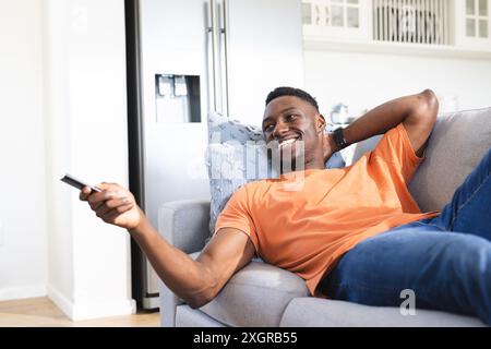 Happy african american man relaxing on sofa watching tv, using remote control. Relaxation, television, technology, entertainment, free time, domestic Stock Photo